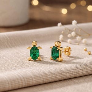 May include: A pair of gold-colored turtle stud earrings with oval-shaped, emerald-green gemstones. The earrings are displayed on a textured, beige surface, with soft lighting and blurred background elements.