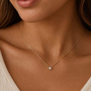 May include: A delicate gold necklace with a small, square-cut diamond pendant. The necklace is shown on a person's neck, highlighting the pendant's sparkle. The fine chain creates a minimalist and refined appearance. The necklace is a piece of jewellery.