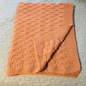 May include: A rectangular knitted blanket in a solid peach color featuring a textured basketweave pattern. The blanket has a folded corner revealing the reverse side of the knit, and it is finished with a simple garter stitch border on all edges.