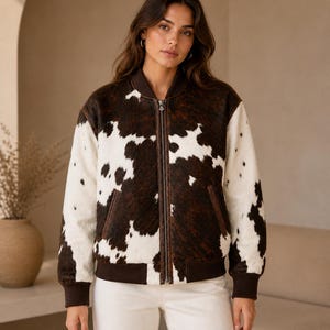 May include: A brown and white cowhide bomber jacket with a zip-up front, brown trim, and two front pockets. The jacket has a ribbed hem and cuffs. The model is wearing white pants.