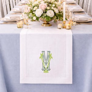 May include: A formal dining table setting with a light blue tablecloth and white runner. The runner features an embroidered green and white monogram. The table is decorated with floral centerpieces, candles, and place settings.