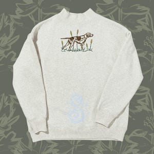 May include: A light gray heathered sweatshirt with a mock neck collar. The center chest features an embroidered graphic of a brown and white spotted hunting dog standing in a field of green grass and tall yellow reeds, rendered in a detailed style.