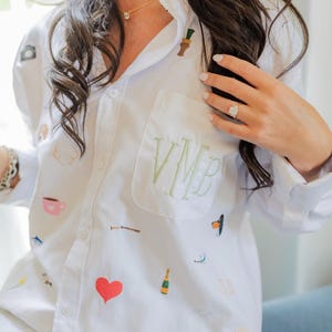 May include: A white button-down shirt featuring a chest pocket embroidered with the green monogram VMB. The fabric is decorated with various small, colorful embroidered icons including a red heart, a yellow sun, a champagne bottle, and a dog.
