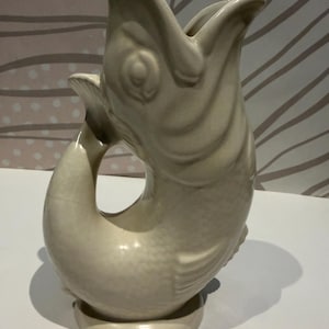 May include: A cream-coloured ceramic jug shaped like a fish with its mouth open at the top. The handle is formed by the curved tail of the fish, which connects to the body. The surface features a textured scale pattern and a smooth glazed finish.