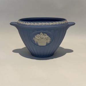 May include: A light blue ceramic bowl featuring a white relief design of a fruit basket in an oval frame. The bowl has a fluted exterior, two small side handles, and a white leaf-patterned border around the top rim, showcasing a matte finish.