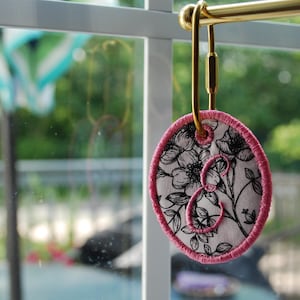 May include: An oval-shaped fabric ornament featuring a black and white floral pattern with a pink embroidered border and a large pink embroidered letter E in the center, suspended from a gold-toned metal carabiner clip on a horizontal rod.
