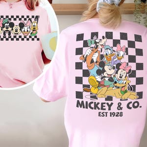 May include: A light pink t-shirt featuring a large graphic on the back of Mickey Mouse, Minnie Mouse, Donald Duck, Daisy Duck, Goofy, and Pluto over a black and white checkered square. The text MICKEY & CO. EST 1928 is printed below the graphic.