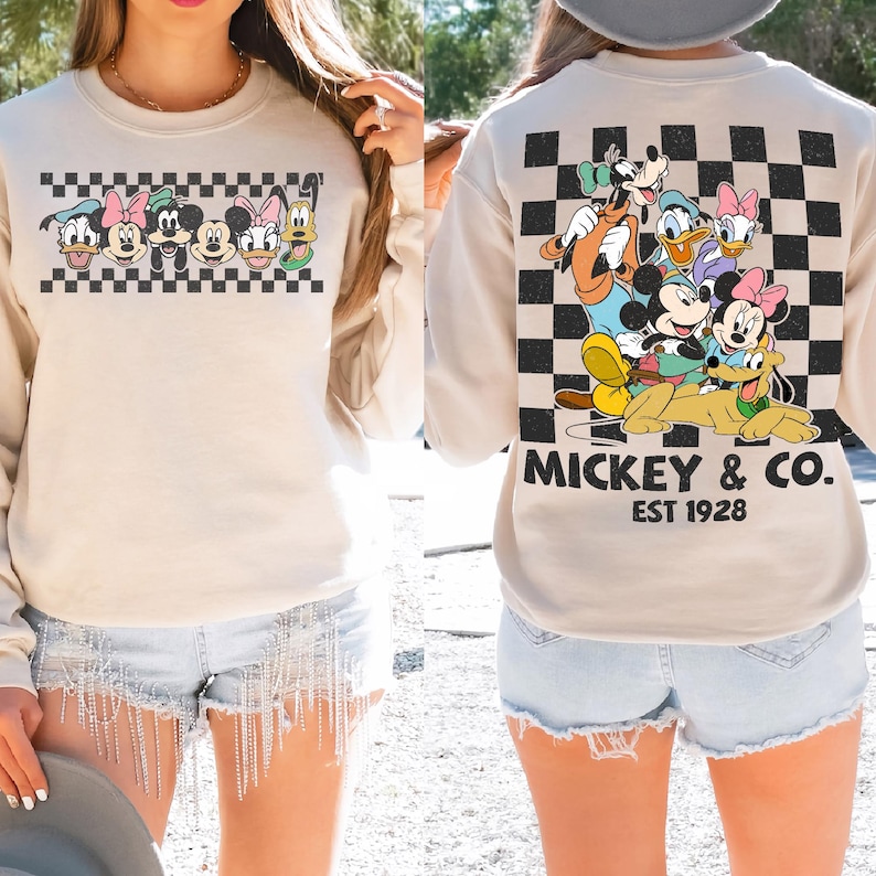 May include: A beige crewneck sweatshirt featuring a graphic of Disney characters on the front and back. The front shows a horizontal checkered strip with character faces. The back displays a large checkered square with the text MICKEY & CO. EST 1928.