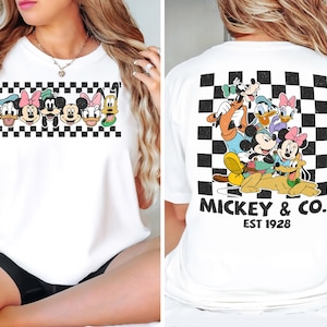 May include: A white t-shirt featuring a graphic print of Disney characters. The front displays a horizontal row of character faces over a black and white checkerboard pattern. The back shows the characters grouped together with the text MICKEY & CO. EST 1928.