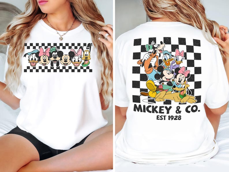 May include: A white t-shirt featuring a graphic print of Disney characters. The front displays a horizontal row of character faces over a black and white checkerboard pattern. The back shows the characters grouped together with the text MICKEY & CO. EST 1928.