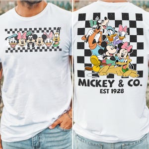 May include: A white t-shirt featuring a graphic of Disney characters on the front and back. The front shows a horizontal checkered strip with character faces. The back displays a large checkered square with characters and the text MICKEY & CO. EST 1928.