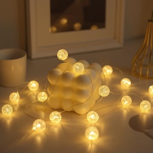 Op de afbeelding: Een reeks warmwitte globe-lampjes is gedrapeerd rond een witte, bubbelvormige kaars. De lampjes zijn verlicht en geven een zachte gloed. Een witte mok en een decoratief gouden metalen object staan op de achtergrond.