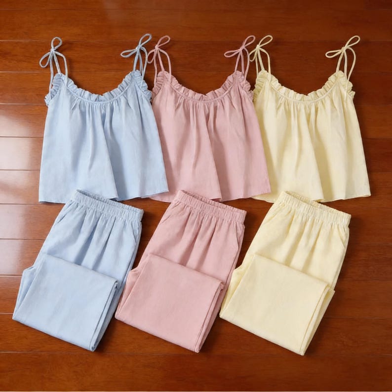Puede incluir: Tres conjuntos de pijama para mujer en colores pastel: azul claro, rosa y amarillo. Cada conjunto incluye un top de camisola con ribete de volantes y tirantes ajustables, combinado con pantalones a juego. Las prendas son de tejido suave y ligero.