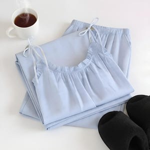 Puede incluir: Conjunto de pijama azul claro con top tipo camisola con volantes y tirantes anudados, combinado con pantalones a juego. Una taza blanca de t&eacute; humeante y zapatillas negras completan la escena.