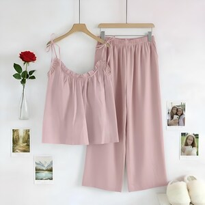 Puede incluir: Un conjunto de pijama rosa claro, que consta de una camiseta sin mangas con tirantes anudados y ribete con volantes, y pantalones de pernera ancha. El conjunto est&aacute; colgado de una percha de madera. Una rosa roja en un jarr&oacute;n y fotos enmarcadas decoran la pared.