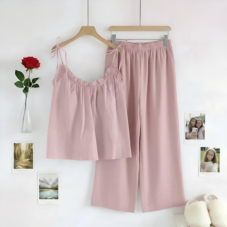 Puede incluir: Un conjunto de pijama rosa claro, que consta de una camiseta sin mangas con tirantes anudados y ribete con volantes, y pantalones de pernera ancha. El conjunto est&aacute; colgado de una percha de madera. Una rosa roja en un jarr&oacute;n y fotos enmarcadas decoran la pared.