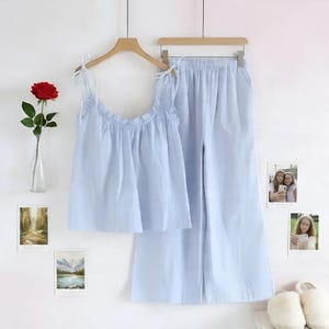 Puede incluir: Un conjunto de pijama azul claro, que consta de una camiseta sin mangas y pantalones de pernera ancha, cuelga de una percha de madera. La parte superior tiene correas anudadas y un escote con volantes. Una rosa roja en un jarr&oacute;n de cristal y fotos enmarcadas decoran la pared.
