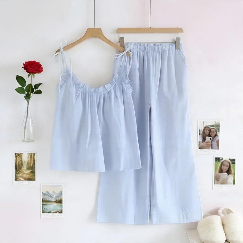 Puede incluir: Un conjunto de pijama azul claro, que consta de una camiseta sin mangas y pantalones de pernera ancha, cuelga de una percha de madera. La parte superior tiene correas anudadas y un escote con volantes. Una rosa roja en un jarr&oacute;n de cristal y fotos enmarcadas decoran la pared.