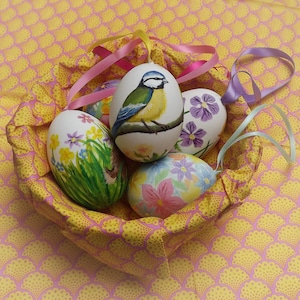 May include: A basket of decorated Easter eggs. The eggs are painted with various designs, including flowers and a blue and yellow bird. The basket is lined with yellow and pink patterned fabric, and ribbons are attached to the eggs.