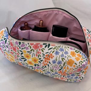 May include: A zipped toiletry bag featuring a white exterior with a colourful floral pattern in shades of yellow, pink, orange, purple, and green. The interior is lined with light pink fabric and contains multiple compartments holding hair tools.
