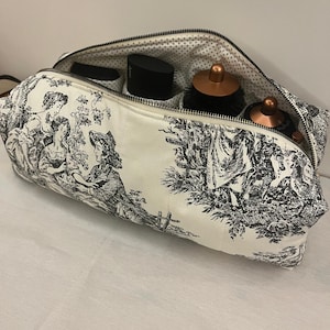 May include: A zipped fabric storage case featuring a black and white toile de jouy pattern with pastoral scenes. The interior is lined with a white fabric featuring small black polka dots and contains several black and copper hair styling tools.
