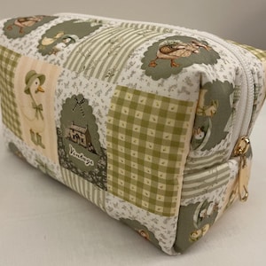 May include: Rectangular cosmetic bag with a patchwork design. The bag features a white zip and a gold-coloured zip pull. The fabric is patterned with geese, houses, and gingham checks in shades of green, cream, and beige.