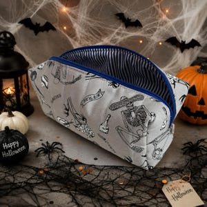 May include: A grey fabric pouch featuring a black and white print of skeletons wearing sombreros, bottles, and the text "Day of the Dead". The pouch has a blue zip closure and an interior lining with a blue and white vertical stripe pattern.