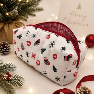 May include: A white quilted zipped pouch featuring a repeating pattern of red and dark green Christmas stockings, ornaments, snowflakes, and pine trees. The interior is lined with a red and white gingham fabric and has a red zip closure.