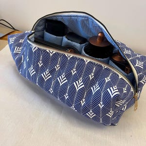 May include: A rectangular fabric toiletry bag featuring a navy blue and white leaf pattern on the exterior and a light blue and white gingham lining. The bag is open with a gold-toned zip and contains several black and copper hair styling tools.