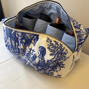 May include: A rectangular toile fabric pouch featuring a blue and white pastoral print with a black zip closure. The interior is lined with a light blue and white gingham fabric and contains several cylindrical compartments for storage.