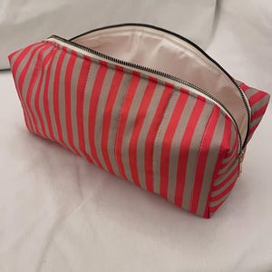May include: A rectangular zipped pouch featuring a vertical striped pattern in vibrant coral red and light beige. The bag has a black zip closure along the top edge and a smooth, solid off-white interior lining visible through the open top.