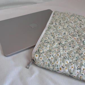 May include: A silver laptop partially inserted into a quilted sage green sleeve featuring a repeating pattern of small white daisies with yellow centres. The sleeve has a white zip closure with a silver metal pull tab along the top edge.