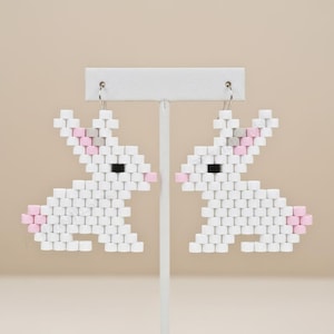 May include: A pair of white bunny earrings made from small, square beads. The earrings feature pink and gray accents for the ears and tail, with black square beads for the eyes. The earrings are displayed on a white earring stand.