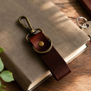 May include: A brown leather keychain with a brass clasp and fittings. The keychain is attached to a brown leather strap with a brass stud. The keychain rests on a grey book. Keys and a brown leather wallet are in the background.