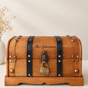 May include: A wooden chest with a curved lid, featuring two vertical black leather-like straps, brass-toned metal studs, and a central brass-toned combination padlock. The front of the chest is engraved with the text The Sylvesters.