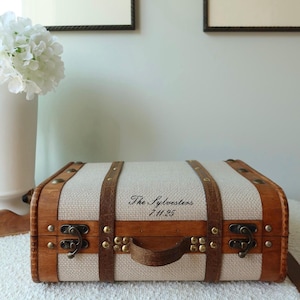 Pu&ograve; includere: Una piccola valigia in stile vintage con corpo in tessuto beige chiaro e struttura in legno. La valigia ha cinghie in pelle marrone, una maniglia e chiusure in metallo. Il testo "The Sylvesters 7.11.25" &egrave; stampato sul davanti.