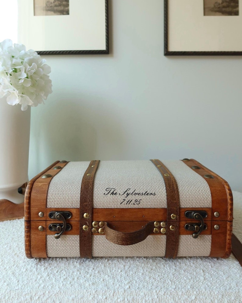 Pu&ograve; includere: Una piccola valigia in stile vintage con corpo in tessuto beige chiaro e struttura in legno. La valigia ha cinghie in pelle marrone, una maniglia e chiusure in metallo. Il testo "The Sylvesters 7.11.25" &egrave; stampato sul davanti.