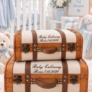 May include: Two rectangular storage trunks with a cream-colored fabric body and brown faux leather trim. Each trunk features metal latches, studs, and a handle. The text 'Baby Calloway Born 7.18.2026' is printed in black on the front of both trunks.