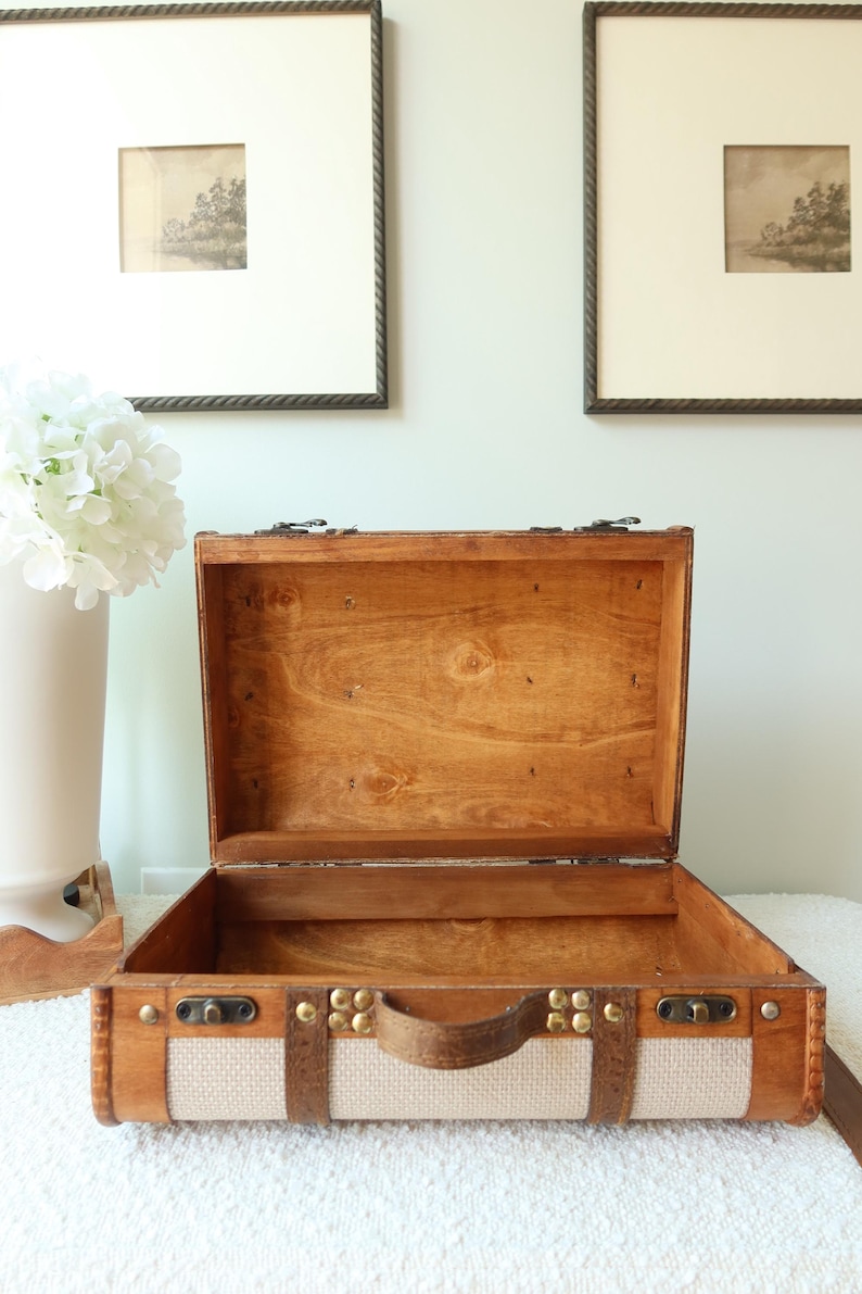 Pu&ograve; includere: Valigia di legno aperta con finitura marrone chiaro, manico in pelle e borchie in ottone. La valigia ha un pannello in tessuto beige sul davanti. Due stampe di paesaggi incorniciate sono appese al muro dietro la valigia.