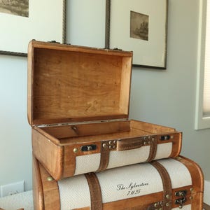 May include: Two vintage-style wooden suitcases stacked on top of each other. The top suitcase is open, revealing the interior. The bottom suitcase has the text "The Sylvesters 7.11.25" printed on it. Both have tan fabric and leather straps.