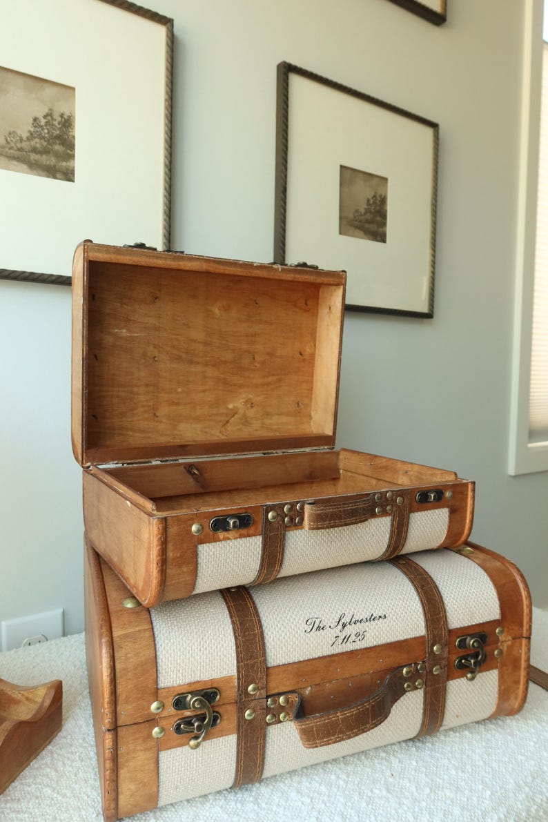 May include: Two vintage-style wooden suitcases stacked on top of each other. The top suitcase is open, revealing the interior. The bottom suitcase has the text "The Sylvesters 7.11.25" printed on it. Both have tan fabric and leather straps.
