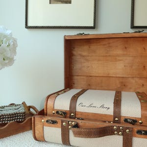 May include: Two wooden decorative trunks with leather straps and brass accents. The top trunk is open, revealing a beige fabric interior. The text "Our Love Story" is printed on the fabric. The trunks are on a white surface.