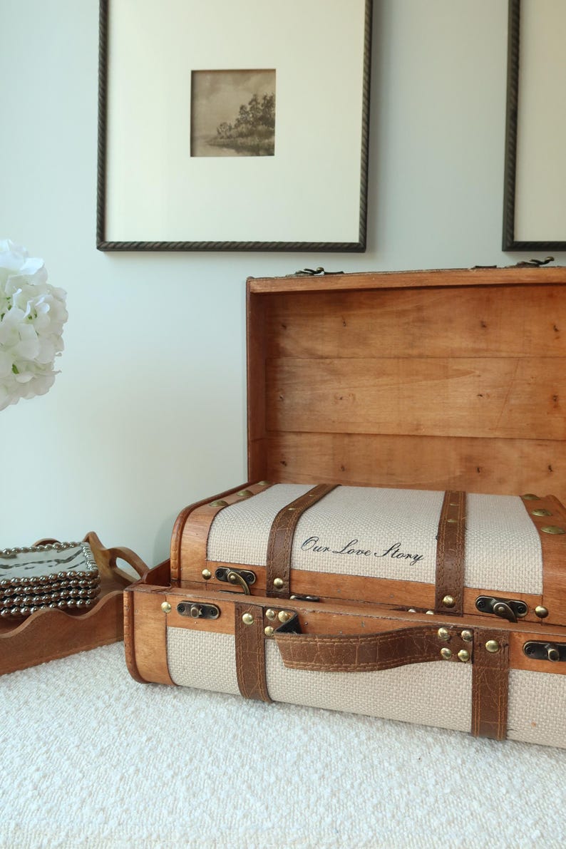 May include: Two wooden decorative trunks with leather straps and brass accents. The top trunk is open, revealing a beige fabric interior. The text "Our Love Story" is printed on the fabric. The trunks are on a white surface.