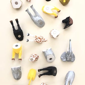 May include: A selection of handmade wooden animal figurines, including a giraffe, elephant, horse, and lion. The figurines are painted in various colours and arranged on a light beige surface. The collection includes a variety of shapes and sizes.