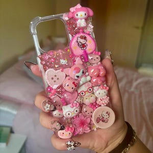 May include: A clear phone case decorated with a variety of pink Hello Kitty charms and embellishments. The case features 3D figures of Hello Kitty, My Melody, hearts, and flowers, creating a playful and decorative accessory.
