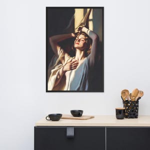 May include: Framed art print featuring a woman in a white garment, bathed in sunlight. The artwork depicts a serene scene with the woman's eyes closed and arms raised. The print is in a black frame, adding a touch of elegance to the artwork.