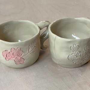 Può includere: Due tazze in ceramica fatte a mano. Una tazza presenta un design floreale rosa, mentre l'altra ha un motivo floreale in rilievo simile. Entrambe le tazze sono di colore beige neutro e hanno una finitura liscia.