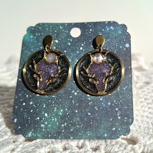 Op de afbeelding: Een paar ronde oorbellen met een goudkleurig metalen frame. Binnenin elke cirkel bevindt zich een zwarte achtergrond met een paarse glitterhertenkop-silhouet, geaccentueerd door een kleine iriserende steen bovenop de kop van het hert.