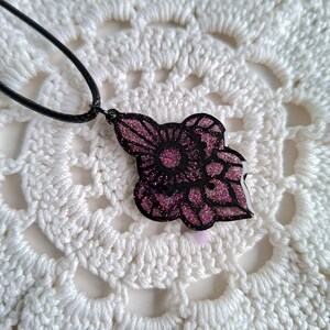 Op de afbeelding: Een zwarte hangerketting met een bloemmotief. De hanger heeft een donkere kleur met een glitterroze bloemenpatroon. De ketting is zwart en de hanger ligt op een wit gehaakt oppervlak.