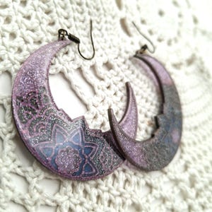 Op de afbeelding: Een paar halve maan oorbellen. De oorbellen hebben een verloop van paars en grijs met glitteraccenten. Elke oorbel heeft een bloemmotief in blauw en zwart. De oorbellen hebben bronzen oorhaken.